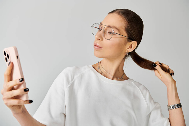 Una joven con una camiseta blanca y gafas sostiene un teléfono celular, sus ojos enfocados en la pantalla. - Foto, Imagen