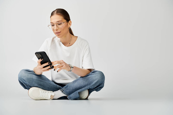 Młoda kobieta w białej koszulce i okularach siedzi na podłodze, pochłonięta telefonem komórkowym.. - Zdjęcie, obraz