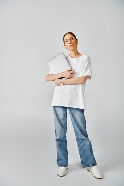 Una joven mujer está de pie con confianza con el ordenador portátil, con una camiseta blanca y gafas sobre un fondo gris. - Foto, Imagen