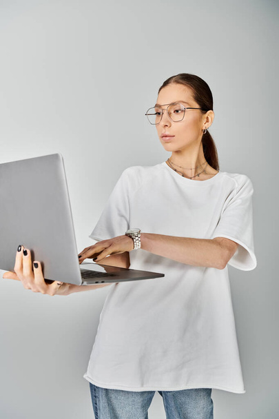 Une jeune femme tient avec confiance un ordinateur portable dans sa main, portant un t-shirt blanc et des lunettes sur un fond gris. - Photo, image