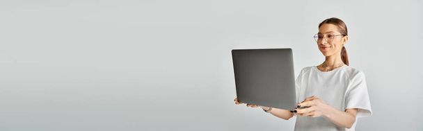 Uma jovem mulher em uma camiseta branca e óculos segurando um laptop em suas mãos contra um fundo cinza. - Foto, Imagem