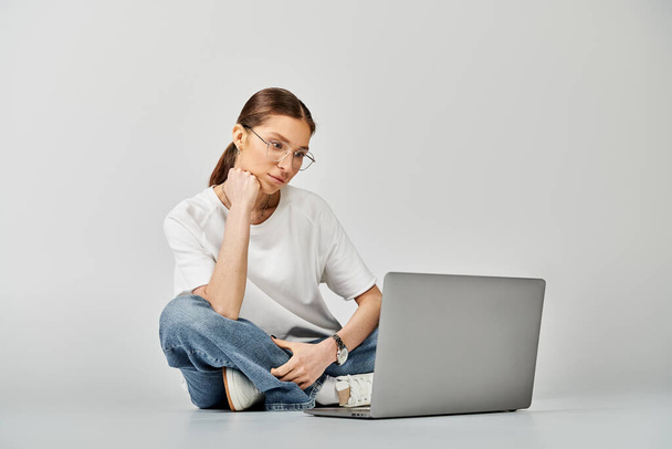 Una joven con una camiseta blanca y gafas se sienta en el suelo, enfocada en la pantalla de su portátil sobre un fondo gris. - Foto, Imagen