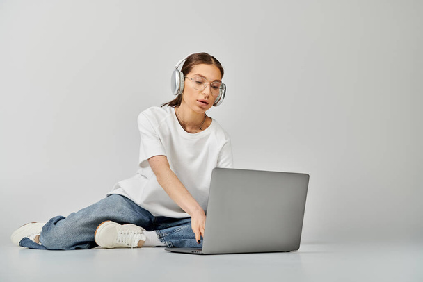 Egy fiatal nő fehér pólóban és szemüvegben ül a padlón, miközben egy laptopon dolgozik.. - Fotó, kép