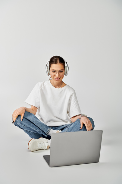 Jeune femme en t-shirt blanc et lunettes se trouve sur le sol avec ordinateur portable, concentré dans les écouteurs. - Photo, image
