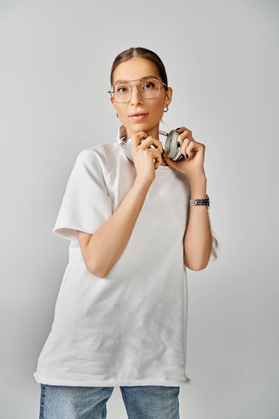 Стильна молода жінка в білій футболці і окулярах тримає навушники на сірому фоні. - Фото, зображення