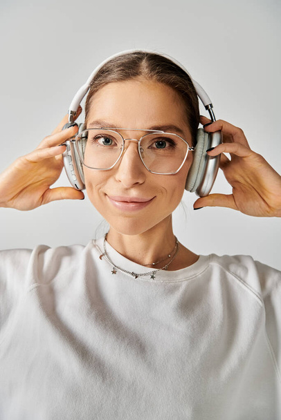 Молодая женщина в очках и наушниках, внимательно слушает на сером фоне. - Фото, изображение