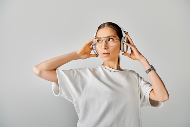 Une femme aux lunettes tient un casque à ses oreilles, exsudant intelligence et sophistication dans un t-shirt blanc sur fond gris. - Photo, image