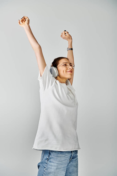 Μια νεαρή γυναίκα με λευκό πουκάμισο και γυαλιά σηκώνει με χαρά τα χέρια της πάνω σε γκρι φόντο.. - Φωτογραφία, εικόνα