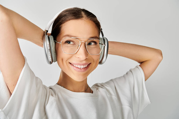 Uma jovem mulher com óculos e fones de ouvido sorri contentemente contra um fundo cinza. - Foto, Imagem