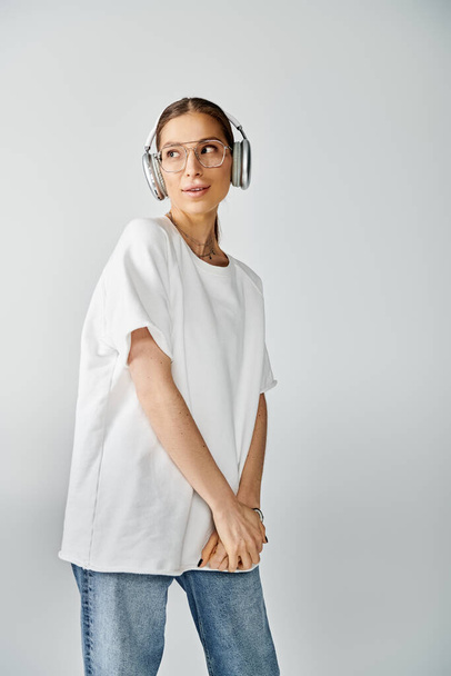 Une jeune femme en t-shirt blanc écoute de la musique à travers des écouteurs sur un fond gris, respirant le calme. - Photo, image