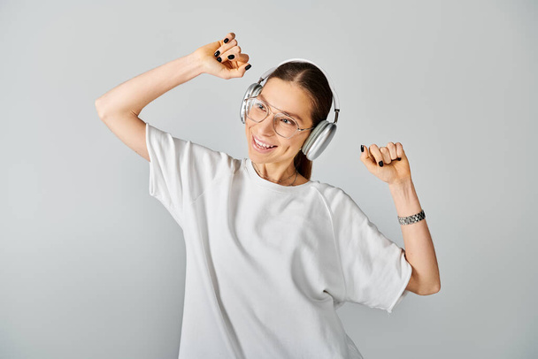 Μια νεαρή γυναίκα με λευκό μπλουζάκι και γυαλιά κρατά ένα ζευγάρι ακουστικά σε γκρι φόντο.. - Φωτογραφία, εικόνα