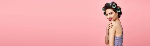 Жінка з бігунками в волоссі, елегантно стоїть на рожевому фоні. - Фото, зображення