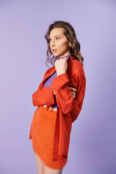 Стильная молодая женщина излучает уверенность, позируя в ярко-оранжевой юбке на фиолетовом фоне. - Фото, изображение
