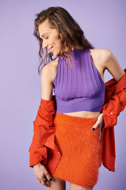 Стильная молодая женщина выделяется в фиолетовой верхней и оранжевой юбке на ярком фиолетовом фоне. - Фото, изображение