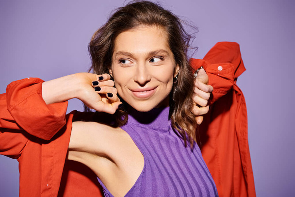 Μια κομψή νεαρή γυναίκα που φοράει ένα μωβ μπλουζάκι κρατά προσεκτικά ένα ζωηρό κόκκινο σακάκι, δημιουργώντας μια εντυπωσιακή αντίθεση χρωμάτων σε ένα μωβ φόντο. - Φωτογραφία, εικόνα