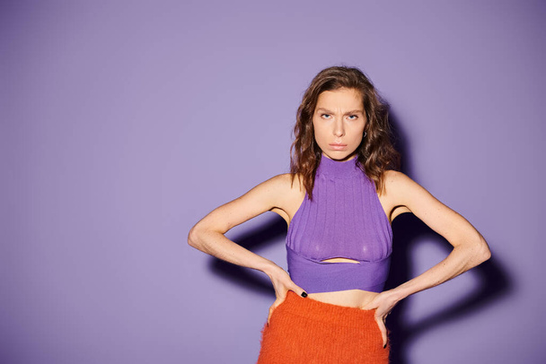 A stylish young woman stands confidently with hands on hips, dressed in vibrant orange attire against a purple background. - Photo, Image