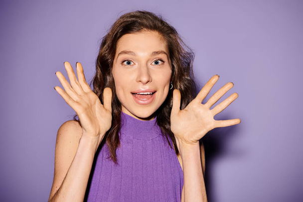 Stylish young woman in a purple top energetically holds her hands up against a vibrant purple background. - Photo, Image