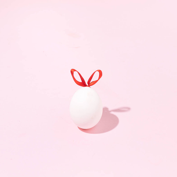 Kırmızı kurdeleli yumurta, minimalist Paskalya tavşanı, pastel pembe arka plana karşı yaratıcı tatil konsepti. Geleneksel bahar dekorasyonu. - Fotoğraf, Görsel