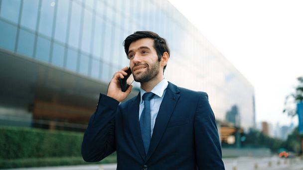 Skilled caucasian project manager calling phone while walking at city with blurred background. Skilled businessman using smart phone talking to project manager with blurred background. Exultant. - Photo, Image