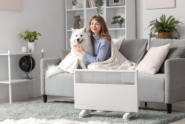 Young woman with Samoyed dog and plaid on sofa warming near radiator at home - Photo, Image