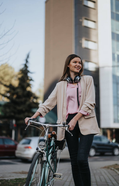 Случайно одетая молодая женщина улыбается стоя с велосипедом на городской улице, наушники вокруг шеи. - Фото, изображение