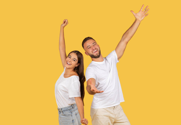 Beyaz bluzlu ve kot pantolonlu neşeli Avrupalı kadın ve erkek kollarını havaya kaldırarak hardal sarısı stüdyo arkaplanına karşı kutlama yapıyorlar. Boş zaman, birlikte eğlence. - Fotoğraf, Görsel