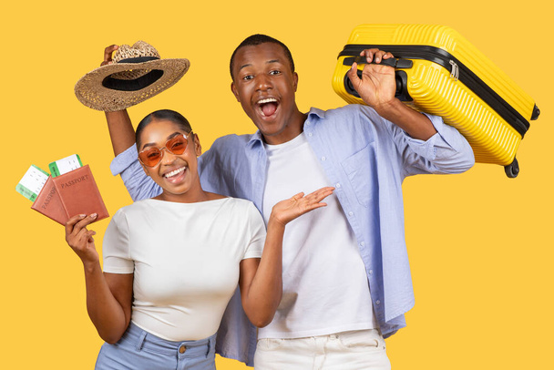 Boldog fiatal fekete pár kezében sárga bőrönd, útlevelek, és szalmakalapos, sugárzó örömmel és várakozással az utazás, sárga háttér - Fotó, kép