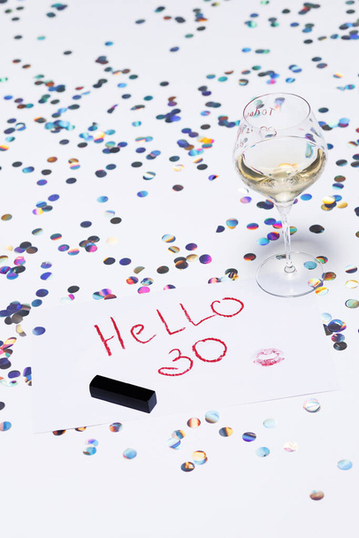 Μια ζωντανή εικόνα συλλαμβάνει την ουσία της στροφής 30, διαθέτει ένα ποτήρι κρασί, πολύχρωμα κομφετί, και ένα χειρόγραφο Hello 30 σημείωση. - Φωτογραφία, εικόνα