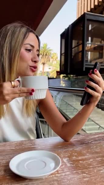 Alkalmi üzleti videó találkozó egy szabadtéri városi kávé: gyönyörű szőke latin ügyvezető kortyolgat kávét, és miután egy videó találkozó a telefonban - Felvétel, videó