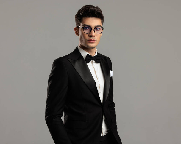 retrato de novio guapo con esmoquin negro y gafas de pie sobre fondo gris con manos en bolsillos - Foto, imagen