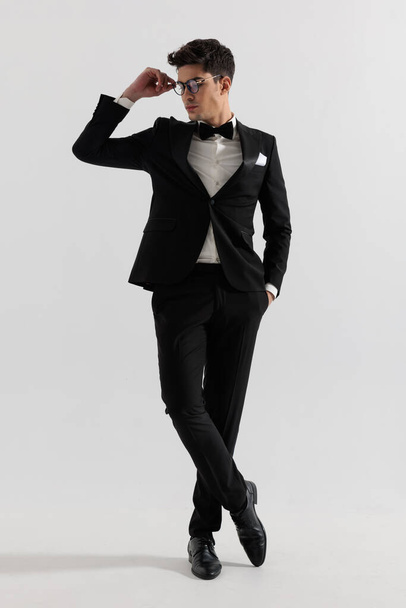 sexy groom in black tux standing with legs crossed and fixing glasses while looking down to side on gray background - Фото, изображение