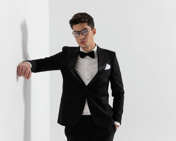 retrato de hombre de moda joven con traje negro y corbata apoyada con el codo contra la pared mientras sostiene el bolsillo sobre fondo gris - Foto, imagen