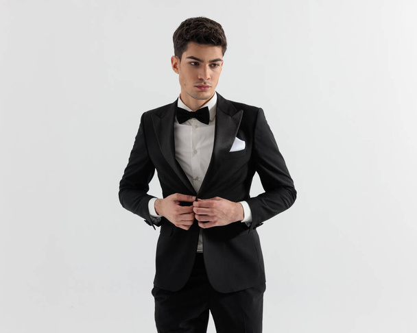 guapo chico modelo buttoning su negro esmoquin mientras mirando hacia abajo a lado en gris fondo - Foto, imagen