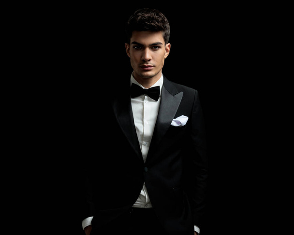 closeup of young man wearing tuxedo and bowtie standing on black background with hands in pockets - Photo, Image