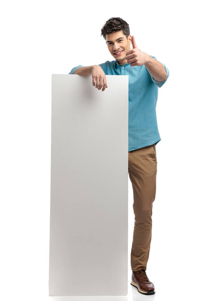 casual man making thumbs up sign while holding billboard and smiling on white background - Photo, Image