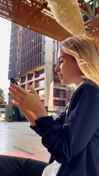 Casual διάλειμμα καφέ σε ένα σύγχρονο δημόσιο σημείο της πόλης: όμορφη και χαλαρή ξανθιά λατίνα εκτελεστικό πίνοντας καφέ και κάνοντας μια βιντεοκλήση με το τηλέφωνο σε μια δημόσια πλατεία - Πλάνα, βίντεο