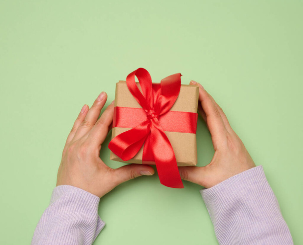 Női kéz kezében egy ajándék doboz csomagolva egy piros selyem szalag a zöld háttér, felső nézet - Fotó, kép