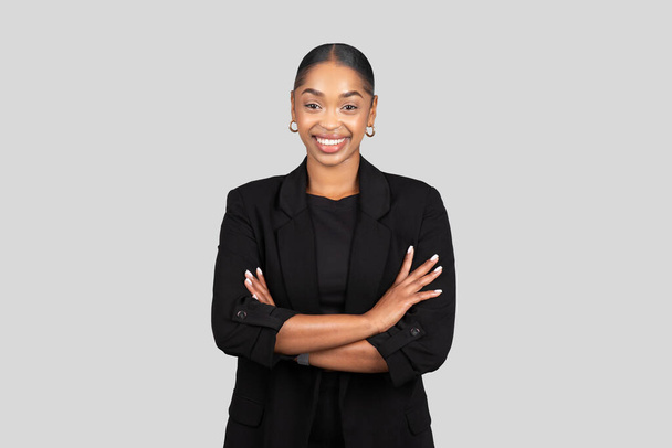 Confident happy African American businesswoman in a sleek black suit stands with arms crossed, smiling assuredly against a neutral gray background, studio. Work, business - Photo, Image