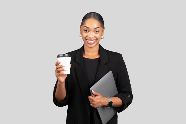 Mujer de negocios afroamericana sonriente sosteniendo una taza de café y una computadora portátil, lista para un día productivo, combinando tecnología y un aumento de cafeína en un traje negro sobre fondo gris, estudio - Foto, imagen