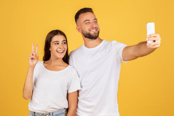 A beaming glad caucasian young woman flashes a peace sign while taking a selfie with a smiling man, capturing a moment of joy and friendship on a sunny yellow background, studio - Photo, Image