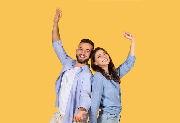 Мужчина и женщина радостно танцуют с высоко поднятыми руками, ярко улыбаясь, в повседневной голубой одежде, празднуя момент на веселом желтом фоне - Фото, изображение