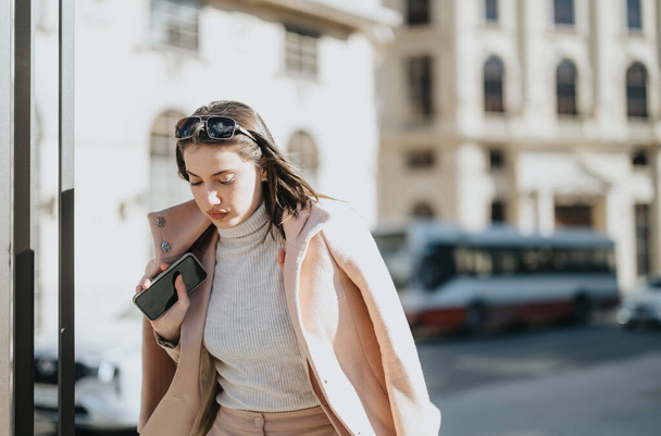 Young businesswoman in a stylish coat engaged with her smart phone. Urban setting with sunlight and buildings in the background. - Photo, Image