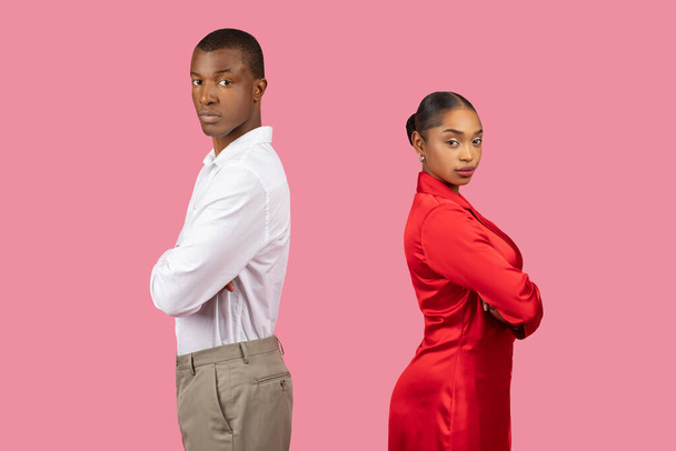 Серьезный черный мужчина и женщина стоят спиной к спине, скрещенные руки, мужчина в белой рубашке и женщина в красном платье, оба излучают сильное присутствие на розовом фоне - Фото, изображение