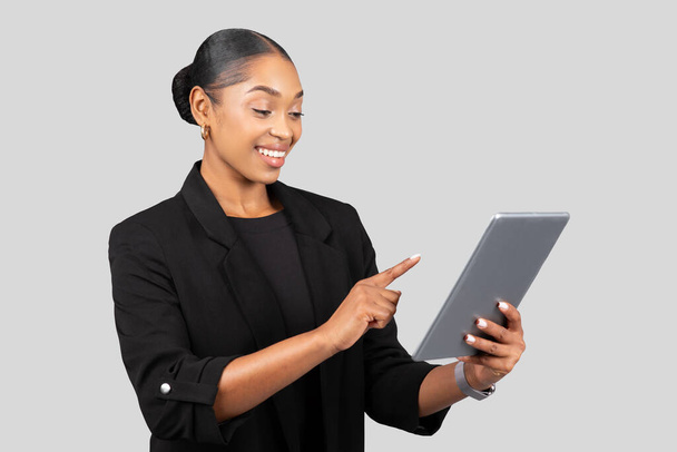 Весела тисячолітня чорна леді в професійному костюмі використовує планшет, вказуючи пальцем, ізольована на сірому фоні студії. Робота, бізнес-додаток, пристрій, спосіб життя у чаті та презентації, стартап - Фото, зображення