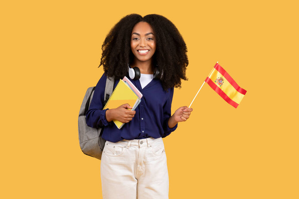 Happy black lady student, wyposażony w plecak i copybooks, dumnie trzyma hiszpańską flagę na żółtym tle, edukacji języka obcego i koncepcji wymiany kulturalnej - Zdjęcie, obraz
