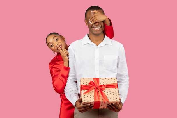 Черная женщина в красном платье игриво закрывает глаза мужчин рукой и держит палец на губах, в то время как парень держит завернутый подарок с красной лентой, передавая сюрприз - Фото, изображение