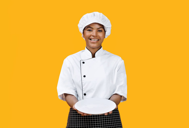 Весела чорна жінка шеф-кухарка з гордістю представляє порожню білу тарілку на камеру, щаслива афроамериканська кулінарна жінка, яка запрошує на смачну їжу, позує на сонячно-жовтому тлі, копіювати простір - Фото, зображення