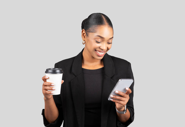 Sorridente donna d'affari afroamericana millenaria multitasking con una tazza di caffè e uno smartphone, godendo di una pausa durante la gestione delle attività di lavoro, isolato su uno sfondo grigio - Foto, immagini