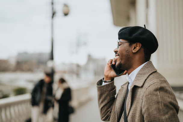Homme d'affaires afro-américain joyeux en tenue élégante et béret ayant un appel téléphonique agréable tout en se tenant debout sur un pont de la ville. - Photo, image