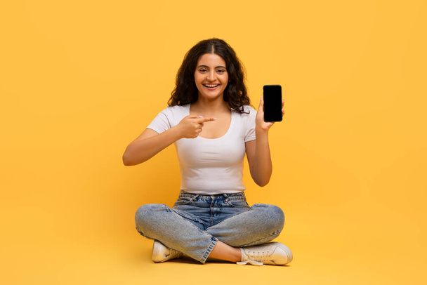 Bom aplicativo móvel educacional, estudar online. alegre atraente encaracolado jovem hindu senhora sentado no chão com smartphone em sua mão, amarelo estúdio fundo - Foto, Imagem